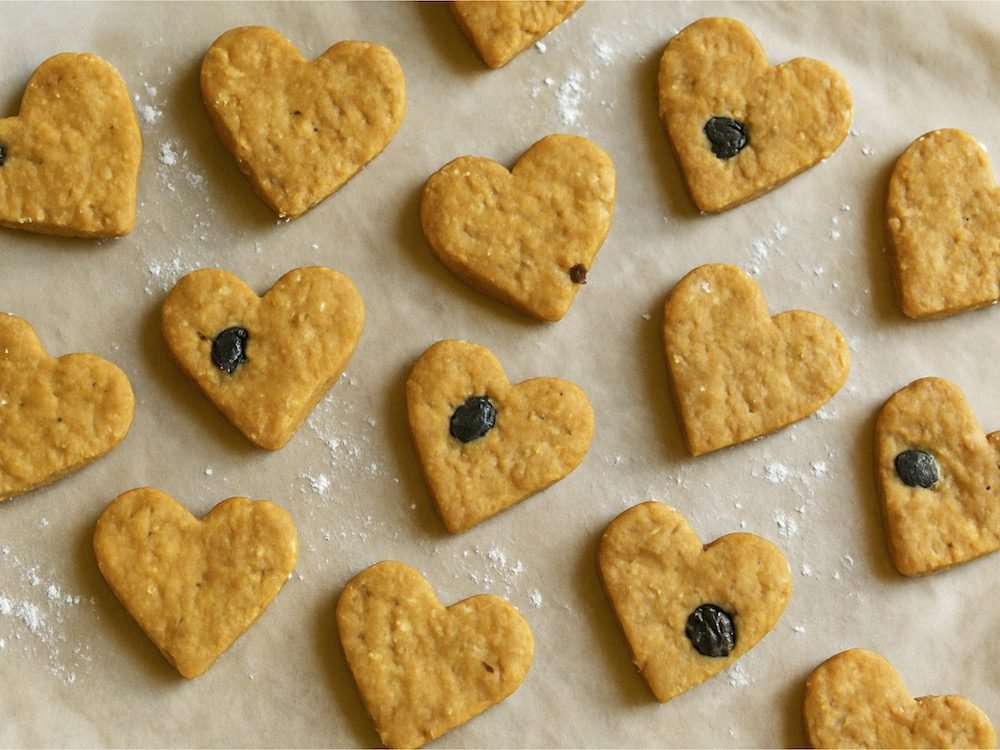 Homemade heart-shaped peanut butter and blueberry dog treats on parchment paper, lightly dusted with flour.
